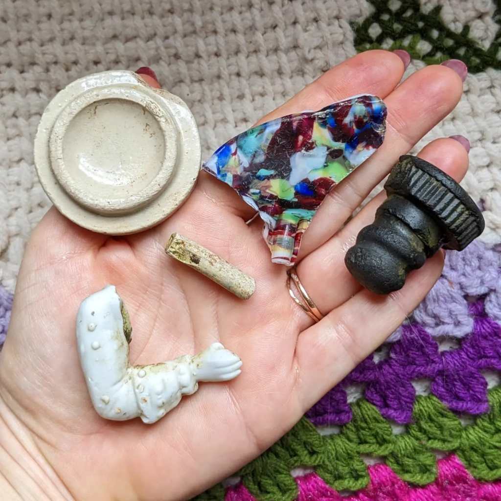 Honor's hand, holding a stoneware bottle top, a piece of carnival glass, a vulcanite twist stopper, a piece of clay pipe stem and the arm of a figurine.