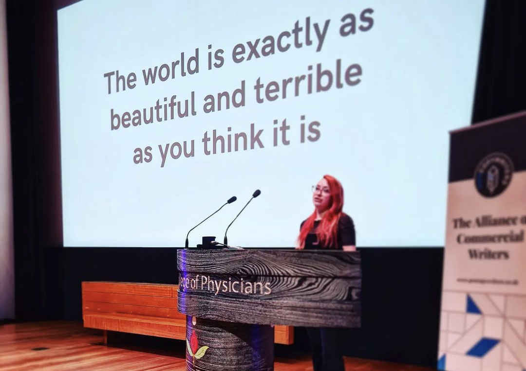 Honor is standing in front of a screen that says 'The world is exactly as beautiful and terrible as you think it is' - it's a talk about neurodivergence and creativity for an audience of copywriters