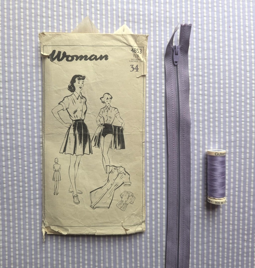 A 1950s sewing pattern, lilac thread and a lilac zip laid on lilac seersucker.