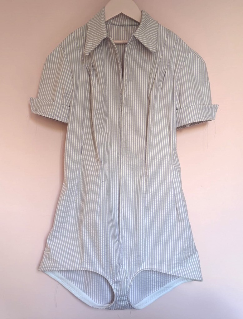 A lilac-striped seersucker gymsuit from a 1950s sewing pattern. It has a dagger collar, big cuffs and it's hanging against a soft pink background.