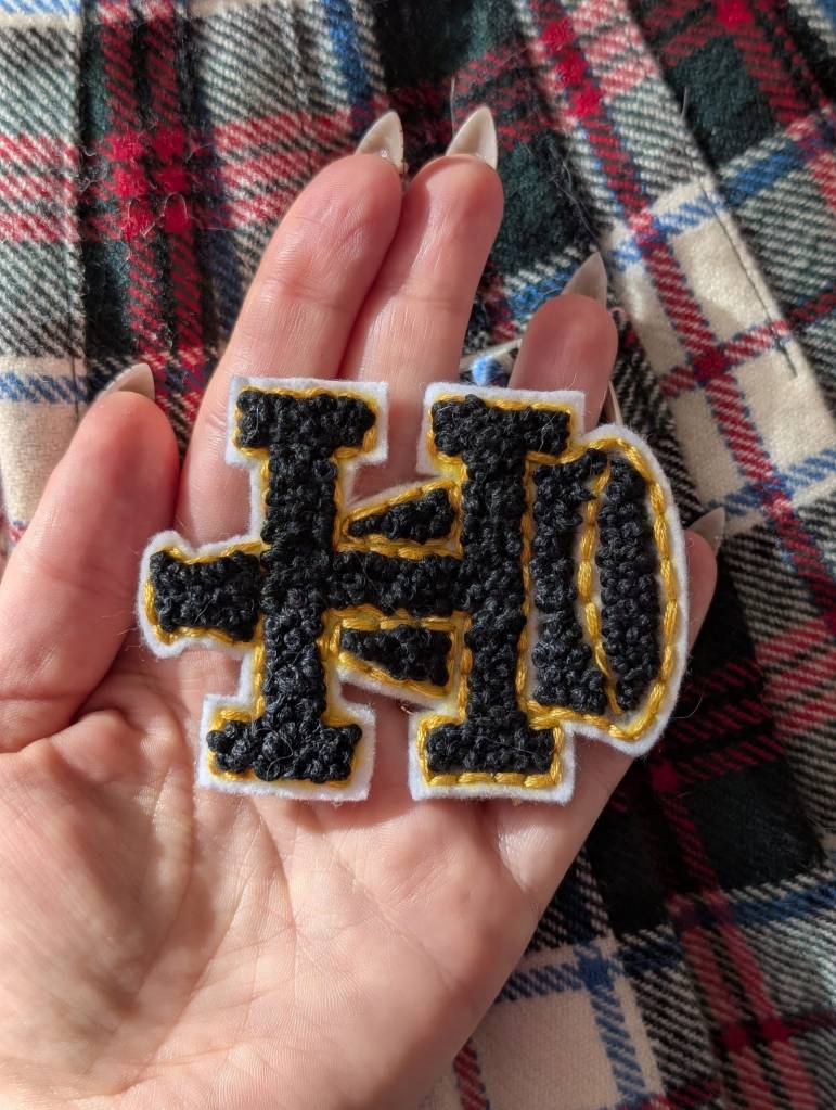 A hand-stitched cheer patch in the shape of an H laid over a megaphone