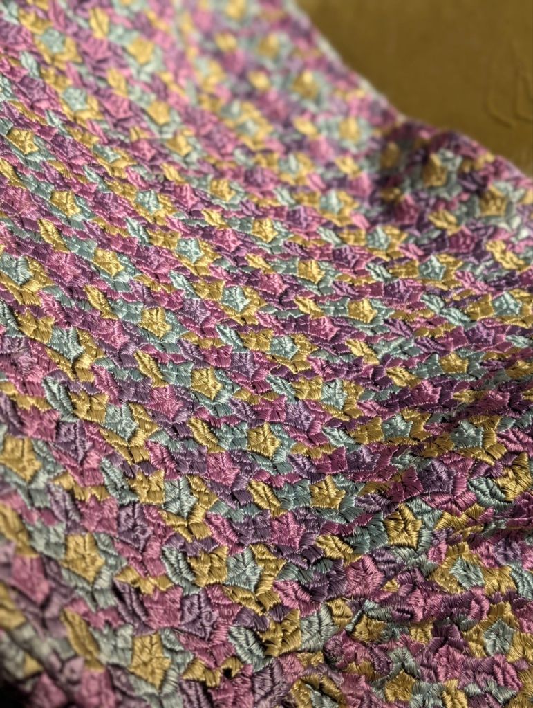 Close-up of a textured fabric featuring a patchwork design in icy blue, olive green, and shades of purple, showcasing intricate leaf patterns.