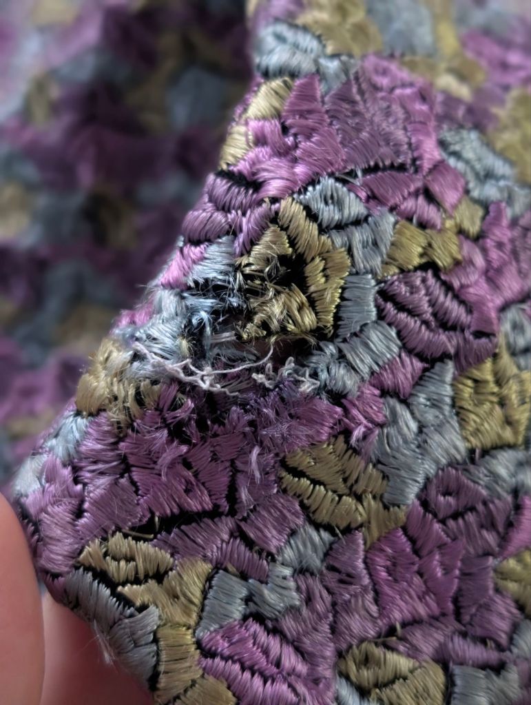 Close-up of a vintage 1930s jacket showing a patchwork of silk-stitched patterns in icy blue, olive green, and purples, featuring a visible hole indicative of wear.