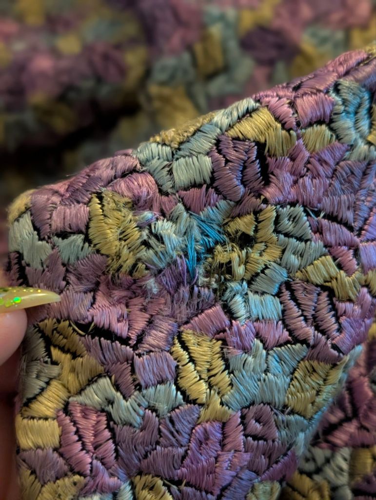 Close-up of a 1930s silk jacket fabric featuring a patchwork of icy blue, olive green, and purple stitched shapes, showing some wear and texture.
