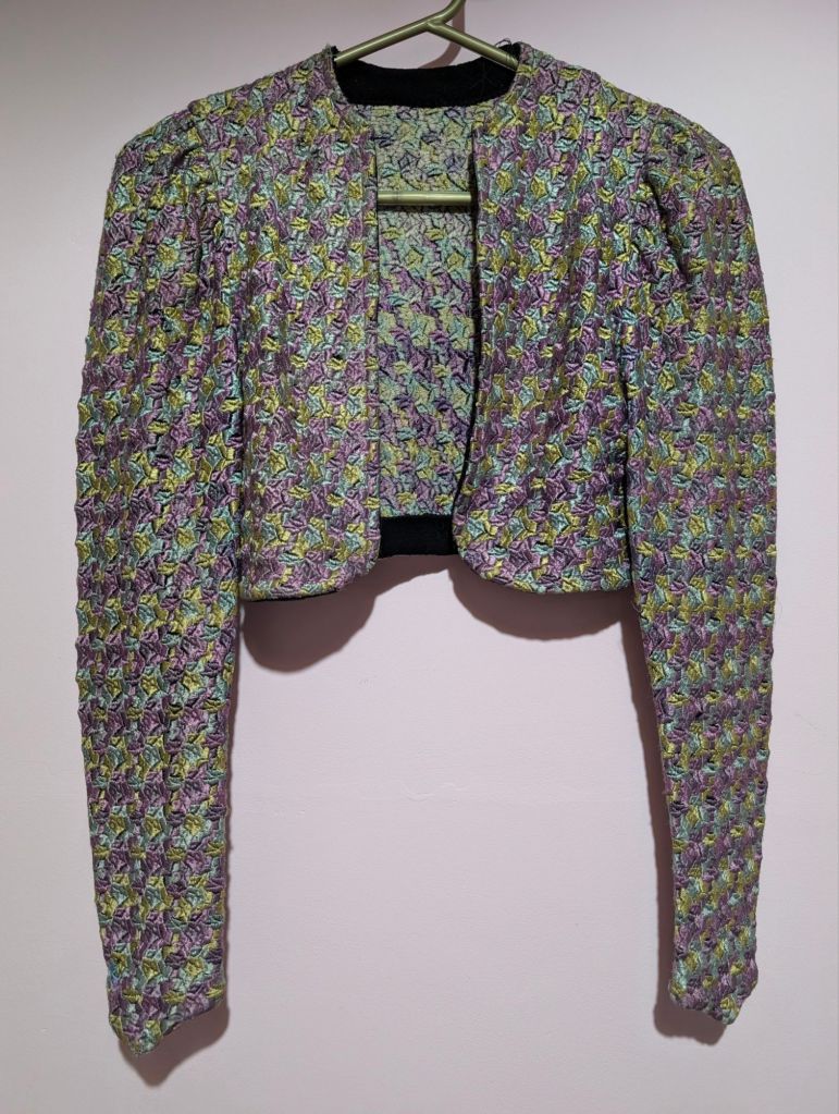 A 1930s cropped jacket made from a patchwork of silk in icy blue, olive green, and purples, displayed on a hanger against a plain background.